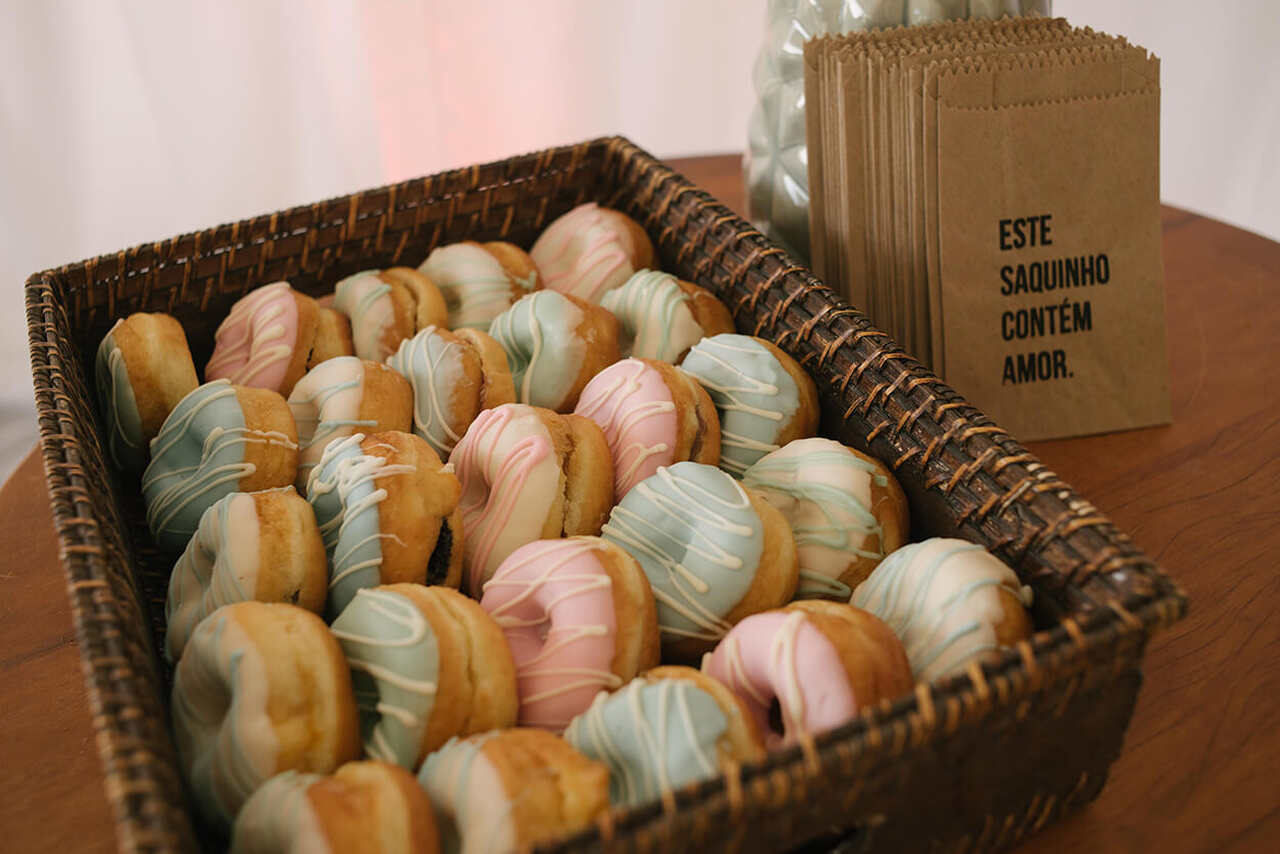 Cesta rústica repleta de donuts decorados com glacê colorido em tons pastel de azul, rosa e branco, acompanhada de saquinhos personalizados com a frase "Este saquinho contém amor".