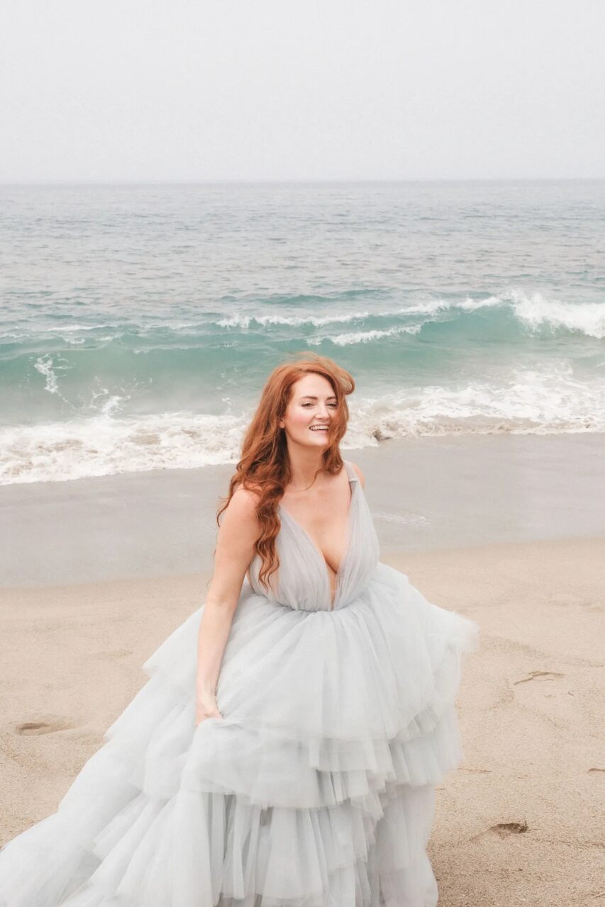 Noiva sorridente na praia, vestida com um volumoso vestido azul-claro em camadas, com o mar ao fundo.