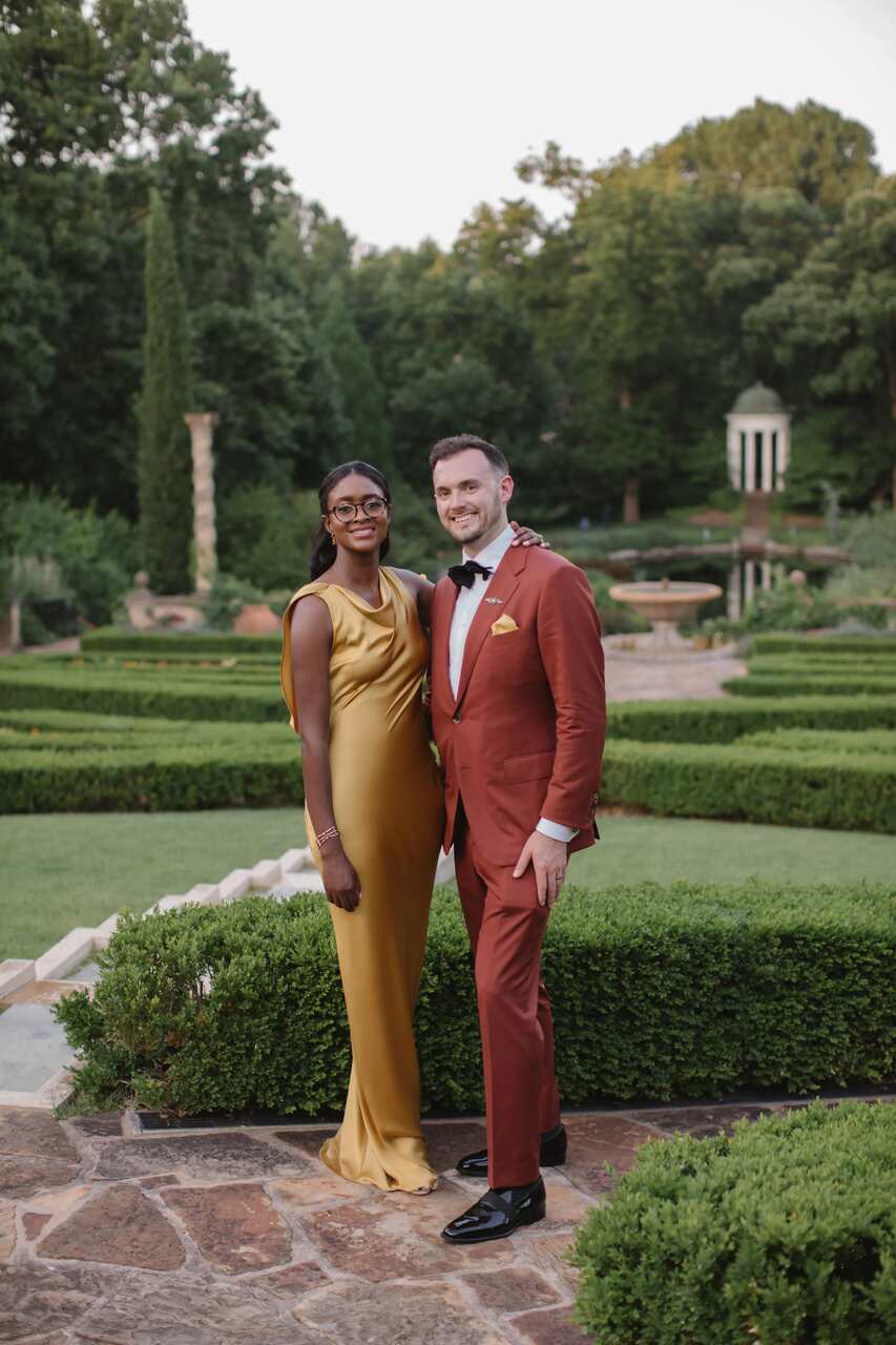 Casal em cenário de jardim; homem em terno marrom e gravata borboleta e mulher com vestido longo dourado elegante.