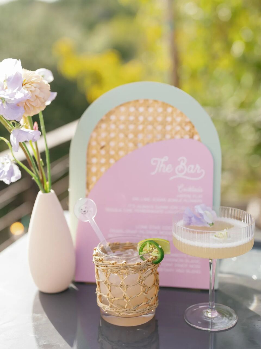 Um close de um bar ao ar livre decorado com um cardápio rosa e verde, ao lado de copos de coquetéis decorados e flores delicadas