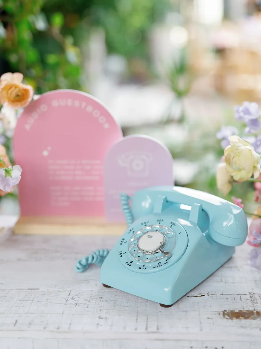 Um telefone retrô azul claro com um display rosa ao lado, indicando um "livro de áudio de convidados" para mensagens de voz personalizadas, cercado por flores.