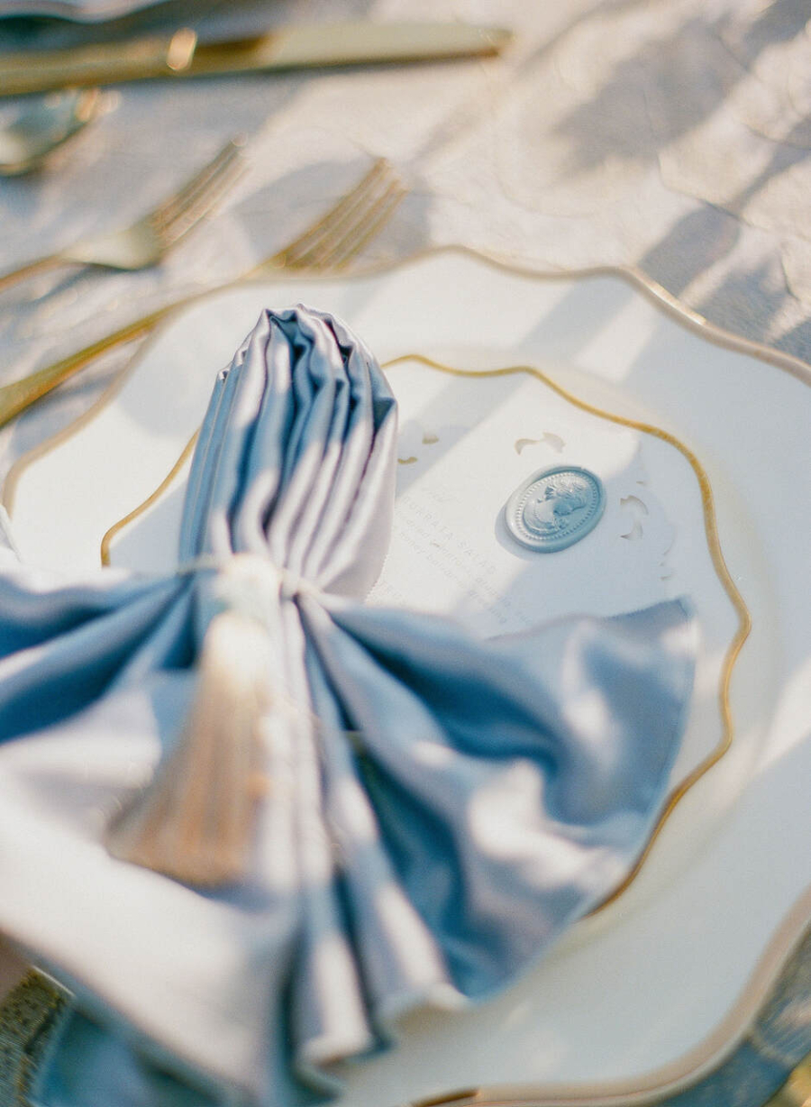 Close-up de um prato de porcelana branco com detalhes dourados em sua borda, sobre uma mesa decorada. No centro do prato, há um guardanapo de tecido azul elegantemente dobrado, preso com um tassel decorativo em tons claros. Ao lado, um pequeno detalhe em relevo cinza que parece ser um selo com uma figura em alto-relevo, adicionando um toque sofisticado à composição. A mesa ao fundo está iluminada por uma luz natural suave, criando um ambiente elegante e refinado.