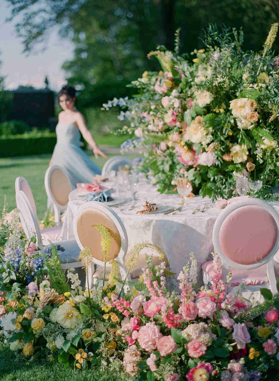 A imagem retrata uma mesa de jantar ao ar livre, cercada por um exuberante arranjo de flores em tons de rosa, amarelo, lilás e branco, complementado por folhagens verdes. A mesa é coberta com uma toalha branca elegante, e as cadeiras têm encostos arredondados com detalhes em rosa. Em segundo plano, uma mulher vestindo um vestido azul claro está levemente desfocada, adicionando um toque de movimento e delicadeza à cena. O ambiente é iluminado por luz natural, destacando a vegetação ao redor e criando uma atmosfera romântica e sofisticada.