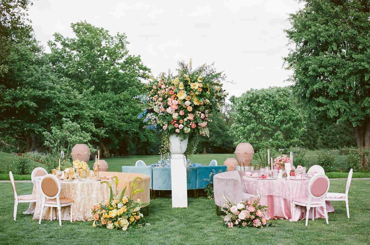 Mesas decoradas em um jardim com tons suaves e arranjos florais vibrantes, destacando uma decoração elegante e natural ao ar livre.