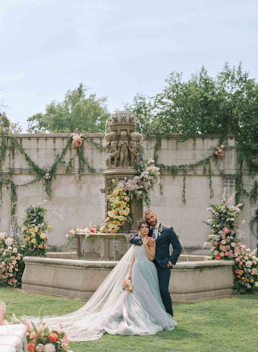 Um casal posa em frente a uma majestosa fonte decorada com flores coloridas, com a noiva usando um vestido azul-claro e um longo véu esvoaçante.