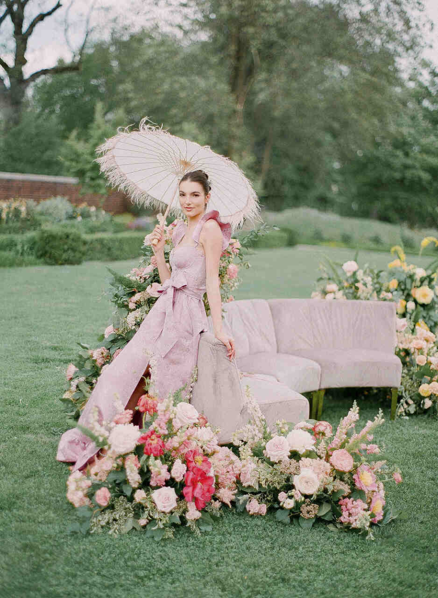 Uma mulher elegante em um vestido lilás posa com um guarda-sol branco em um jardim vibrante, rodeada por flores exuberantes e um sofá aveludado.