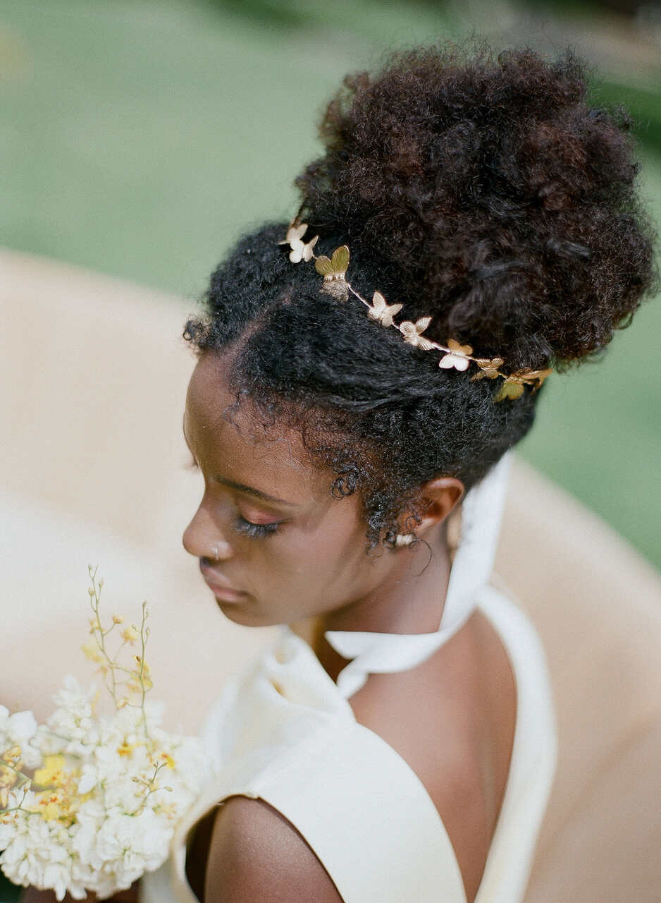 Uma noiva com cabelo afro natural preso em um coque alto, decorado com uma delicada tiara dourada de borboletas. Ela segura um pequeno buquê amarelo.