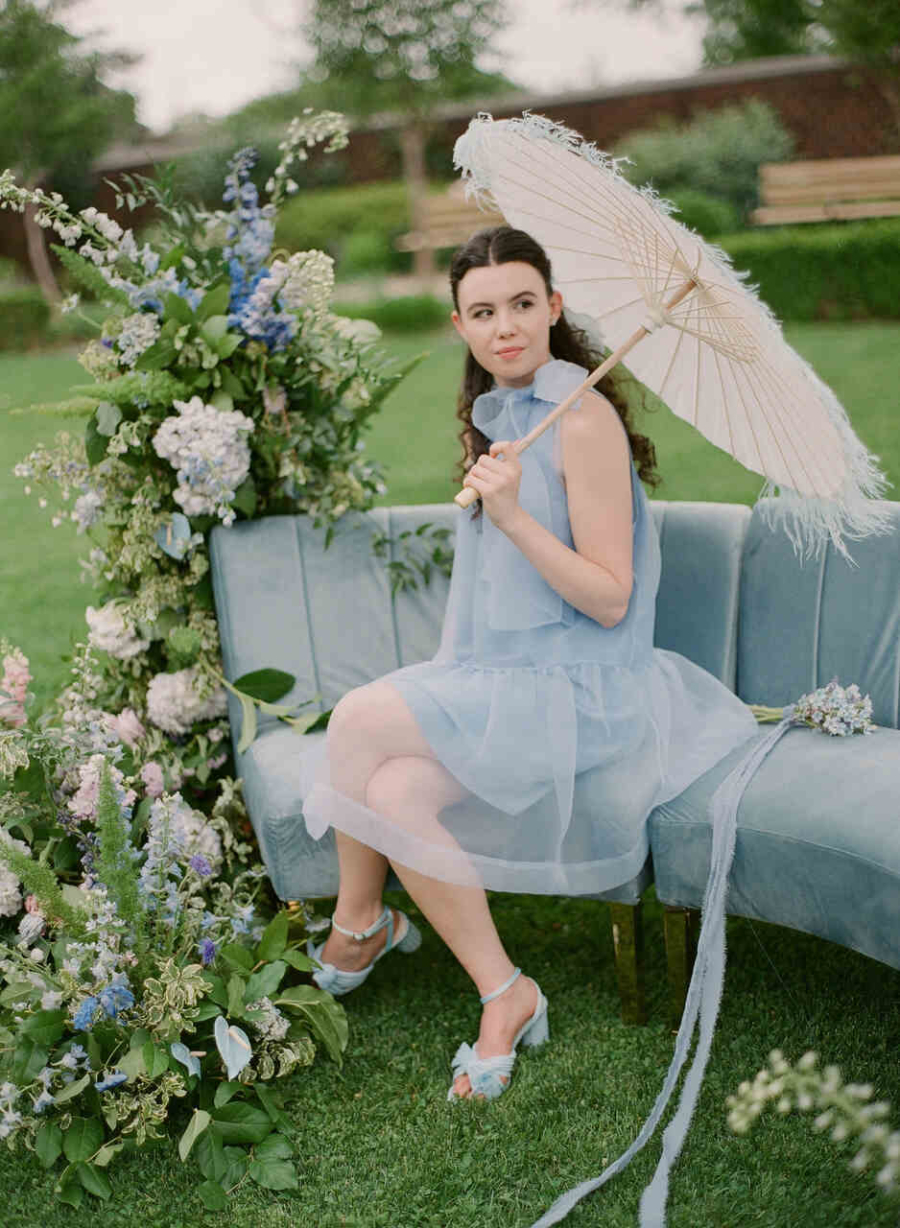 Uma mulher graciosa sentada em um sofá azul-claro no jardim, segura um guarda-sol branco enquanto está cercada por arranjos florais delicados em tons de azul e verde.