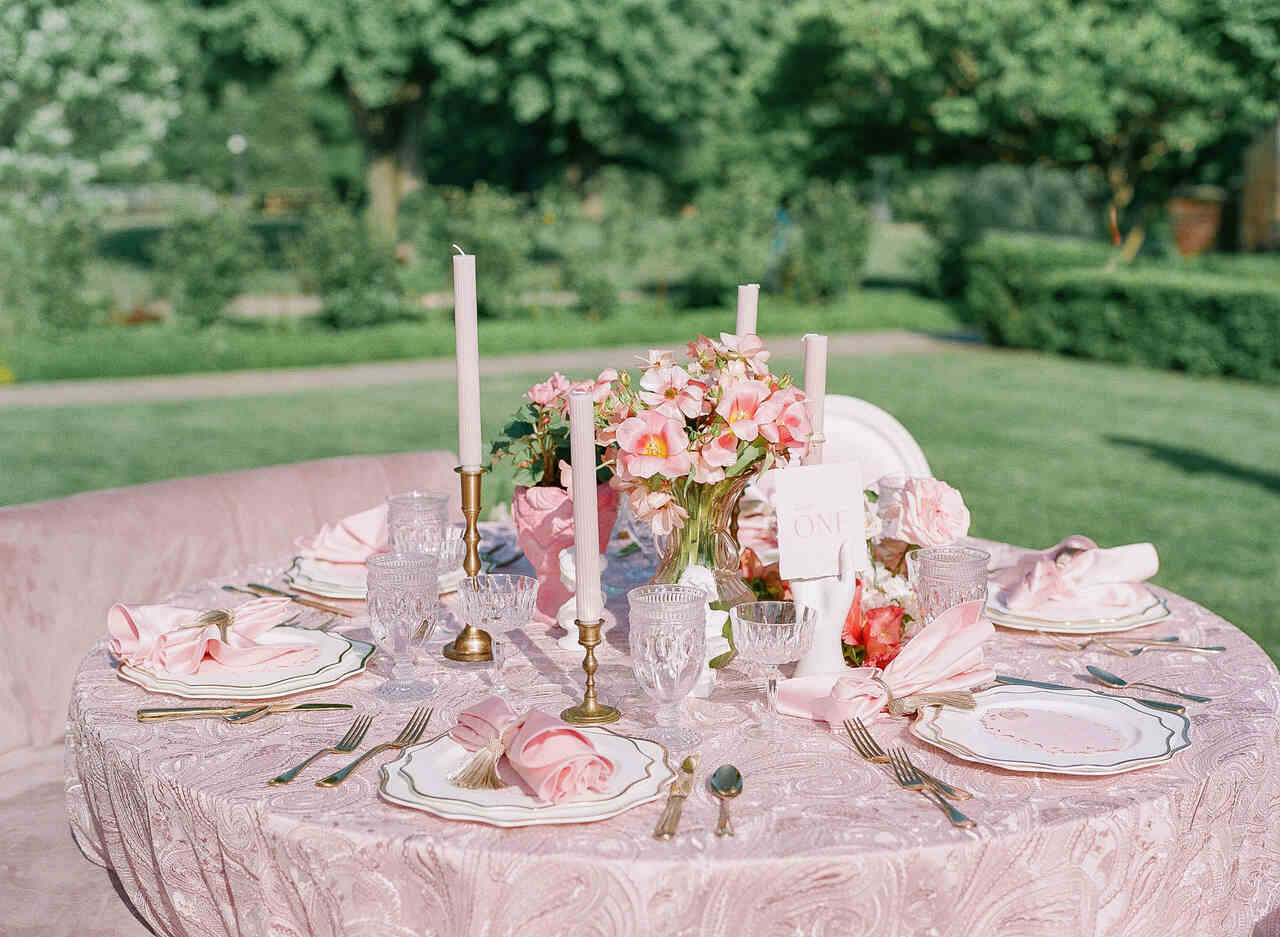 Mesa redonda ao ar livre decorada com uma toalha rosa texturizada, talheres dourados, louça elegante, velas brancas altas e um arranjo floral rosa delicado.