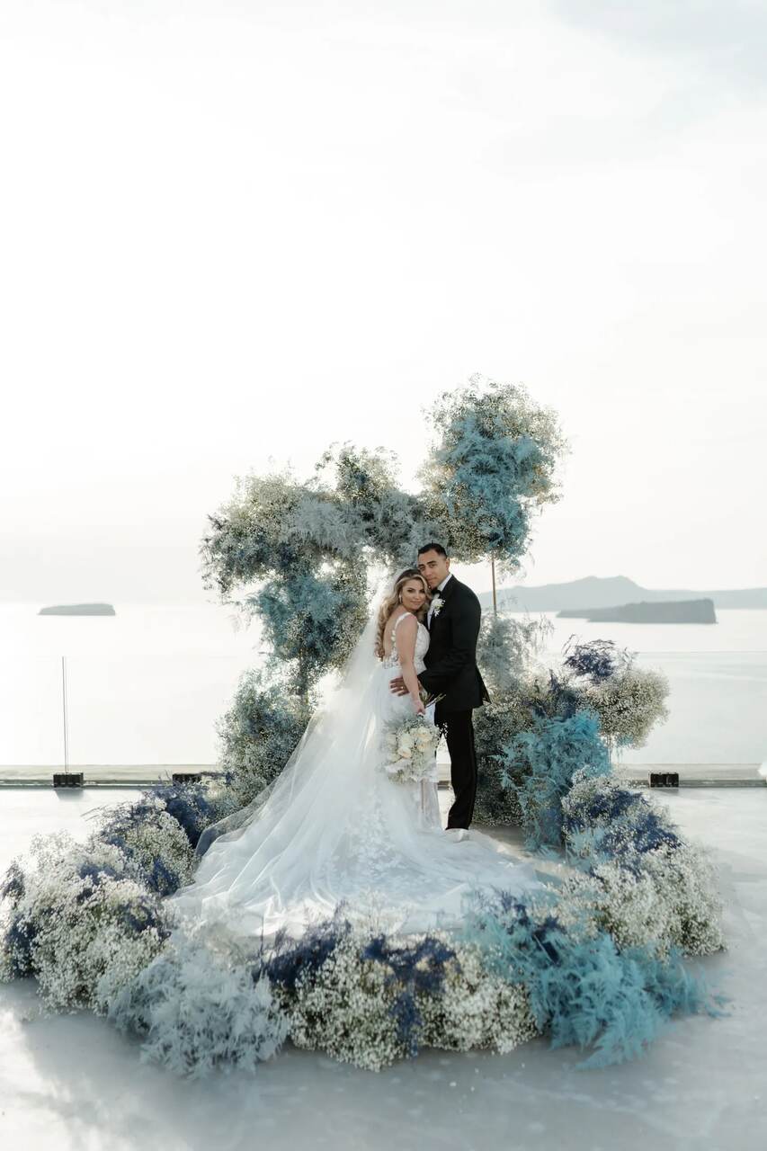 Casal de noivos em um cenário com decoração em tons de azul e branco, destacando flores delicadas e vista para o mar ao fundo.