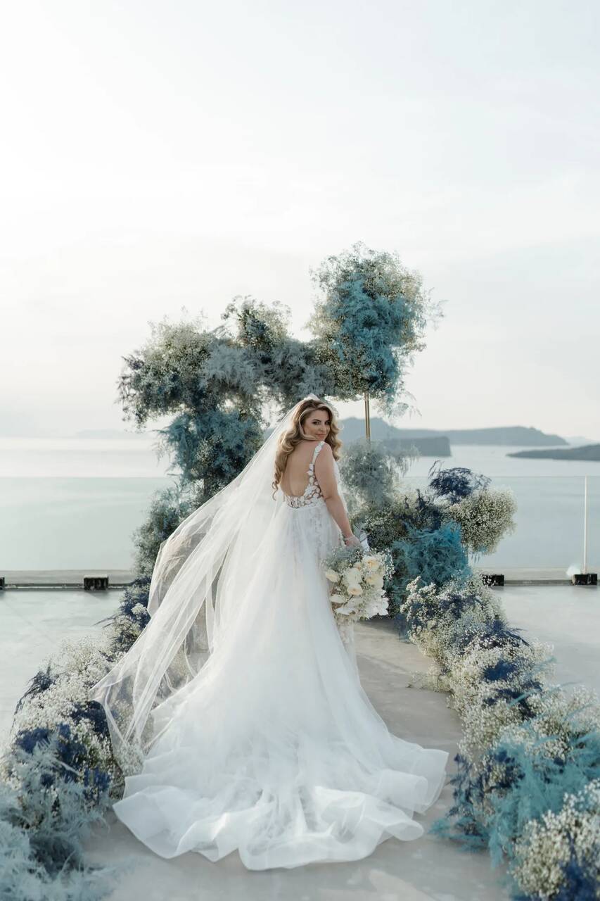 Noiva em um altar à beira-mar, usando um vestido branco com detalhes de pedrarias nas costas e véu fluído, rodeada por uma decoração de flores azuladas.