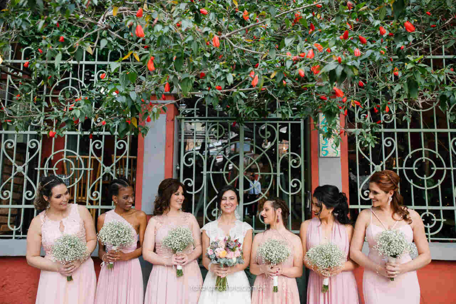 Grupo de madrinhas com vestidos rosados segurando buquês de mosquitinho, ao lado da noiva sorridente.