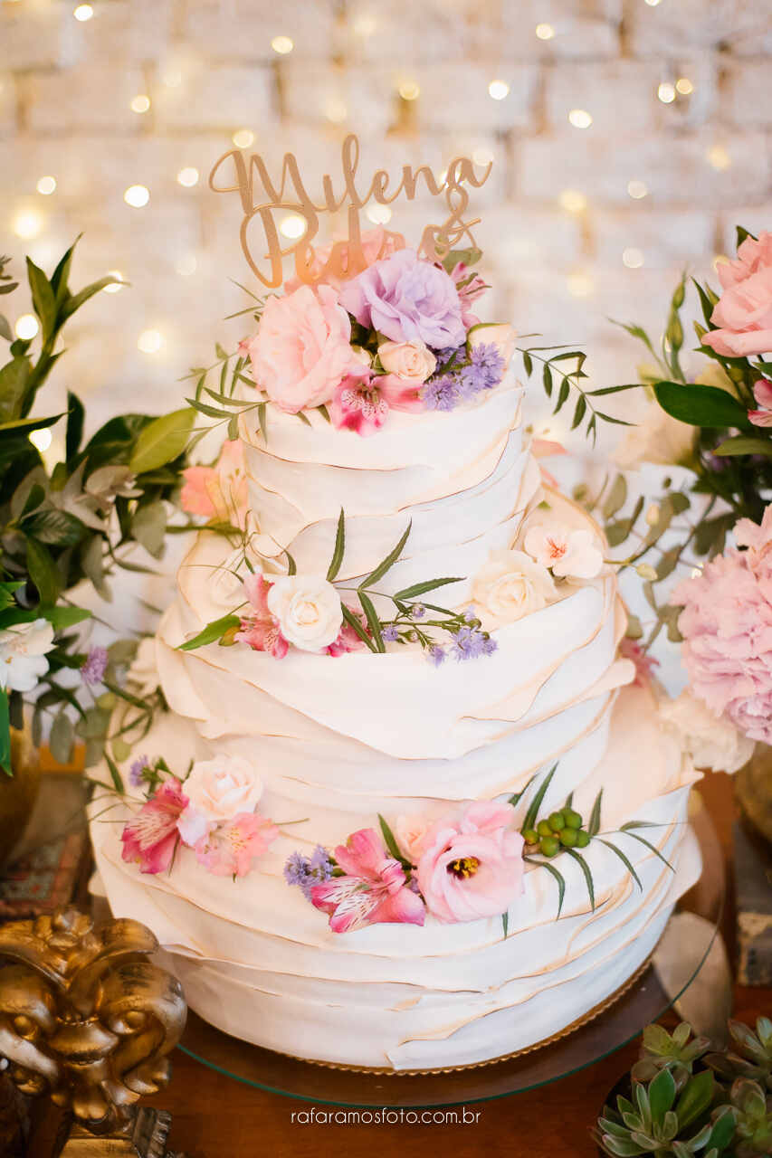 Bolo de casamento branco de três andares com bordas douradas sutis, decorado com flores naturais em tons de rosa, lilás e branco, com o topo personalizado com o nome "Milena e Pedro."