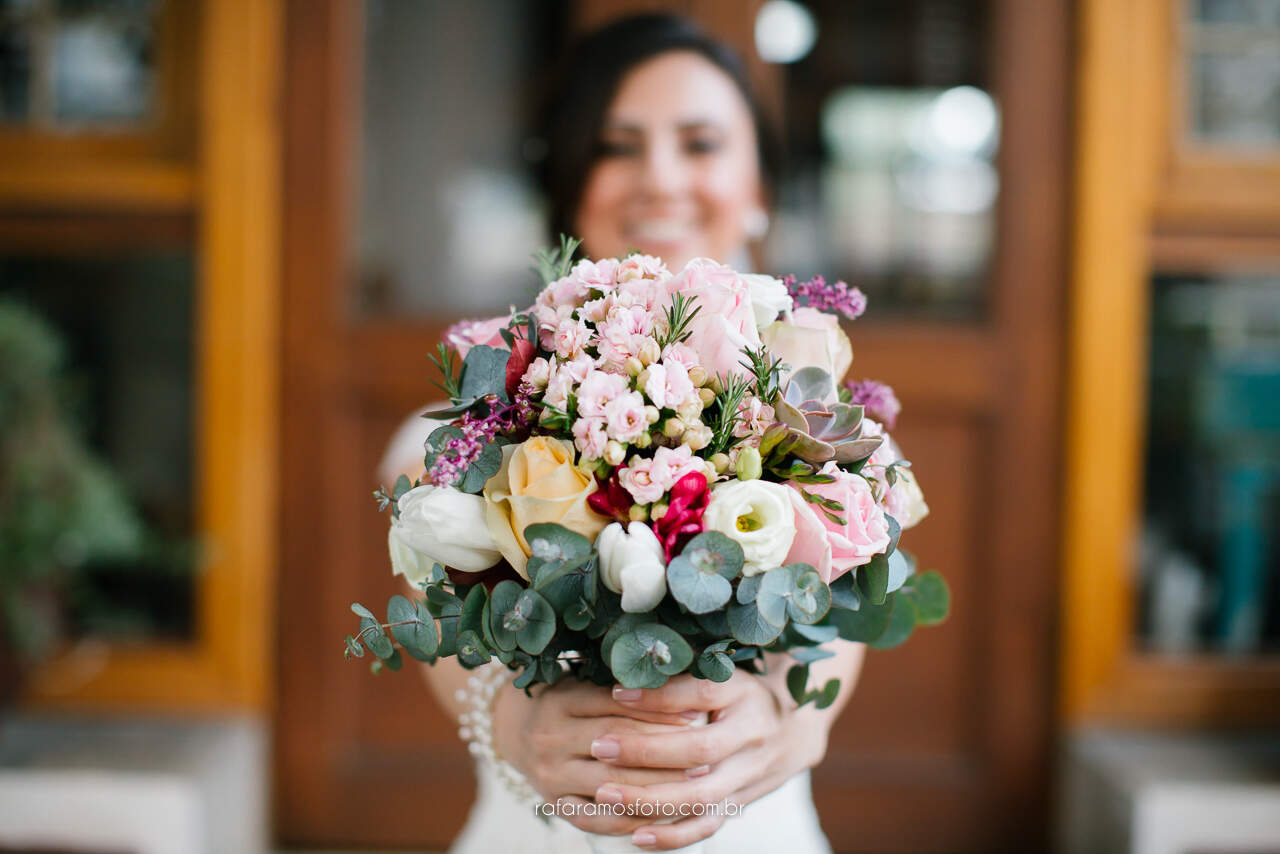 Uma noiva sorridente em segundo plano segura um buquê vibrante com flores em tons de rosa, branco e amarelo, complementado por folhas de eucalipto.