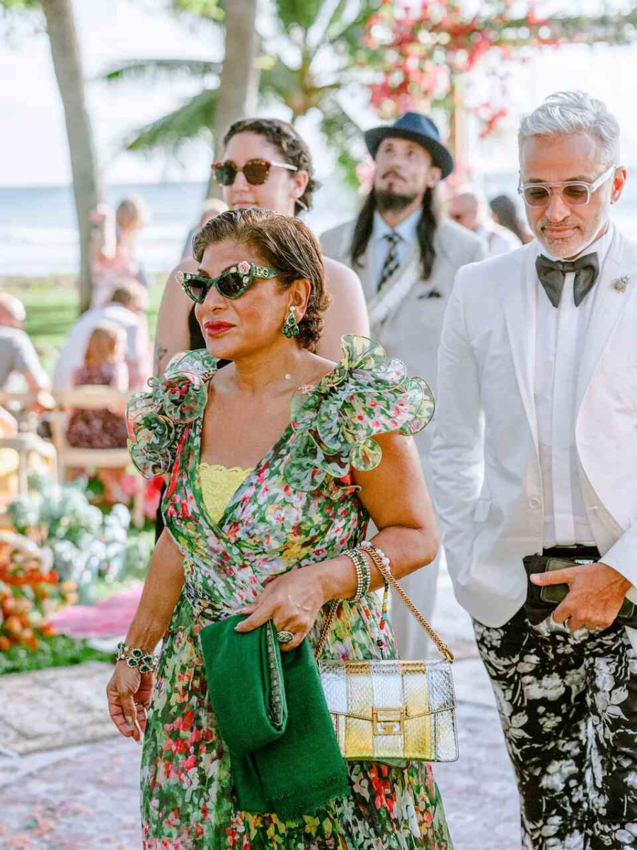 Convidados estilosos em um casamento ao ar livre, incluindo uma mulher com vestido floral e óculos escuros e um homem de terno branco com detalhes modernos