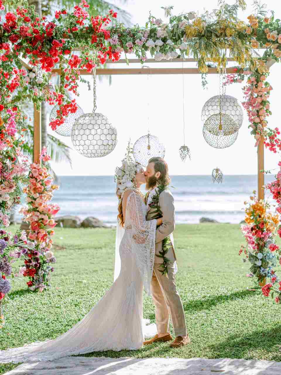 Casal trocando votos sob um arco floral tropical exuberante, com vista para o mar e decoração em tons vibrantes de rosa e branco, em um casamento ao ar livre