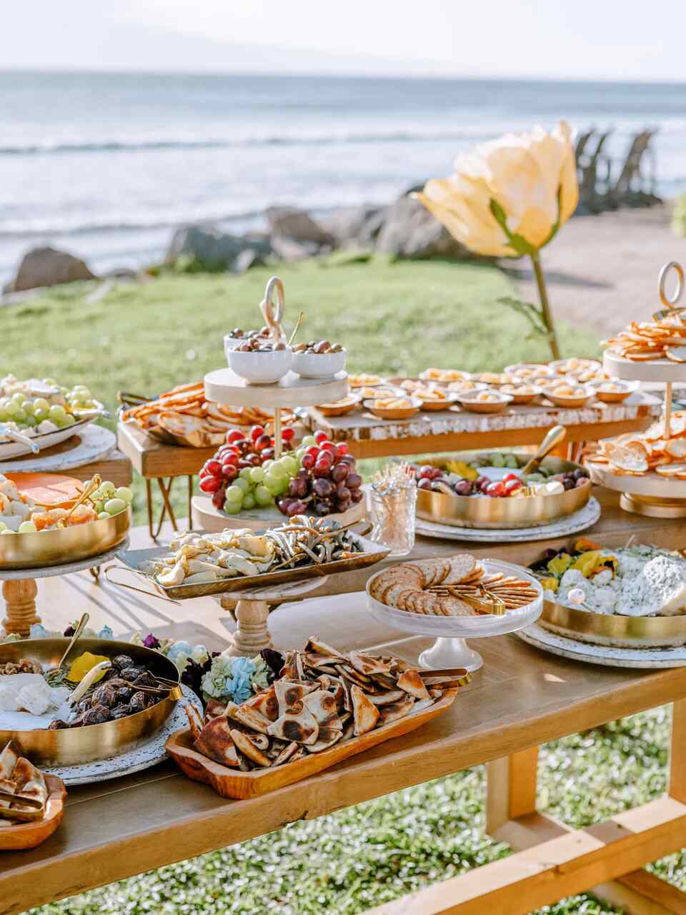 Mesa ao ar livre com uma variedade de queijos, frutas e petiscos dispostos em bandejas elegantes.