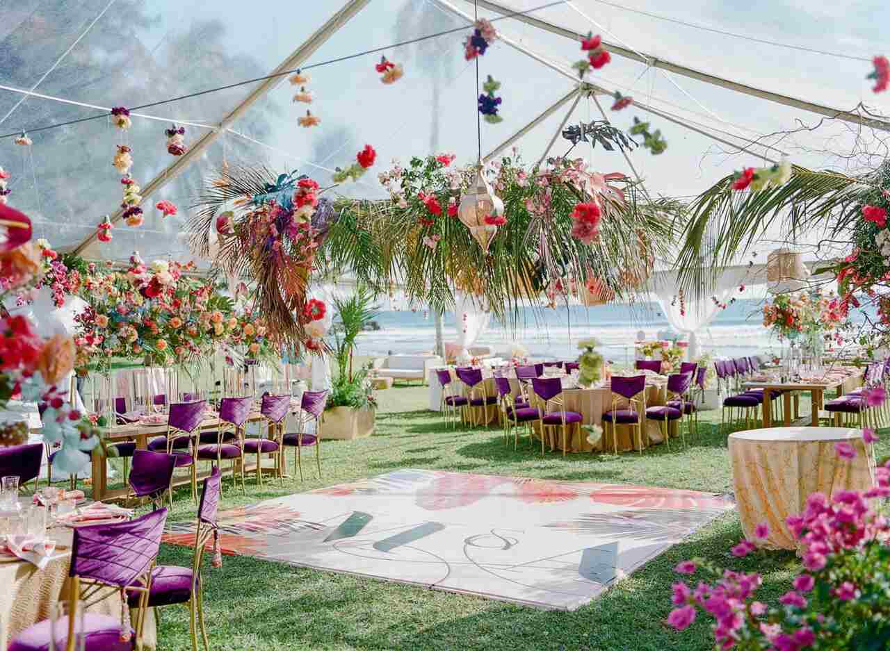 Ambiente da festa de casamento ao ar livre, com mesas e cadeiras na cor roxa, arranjos florais e folhagens suspensos