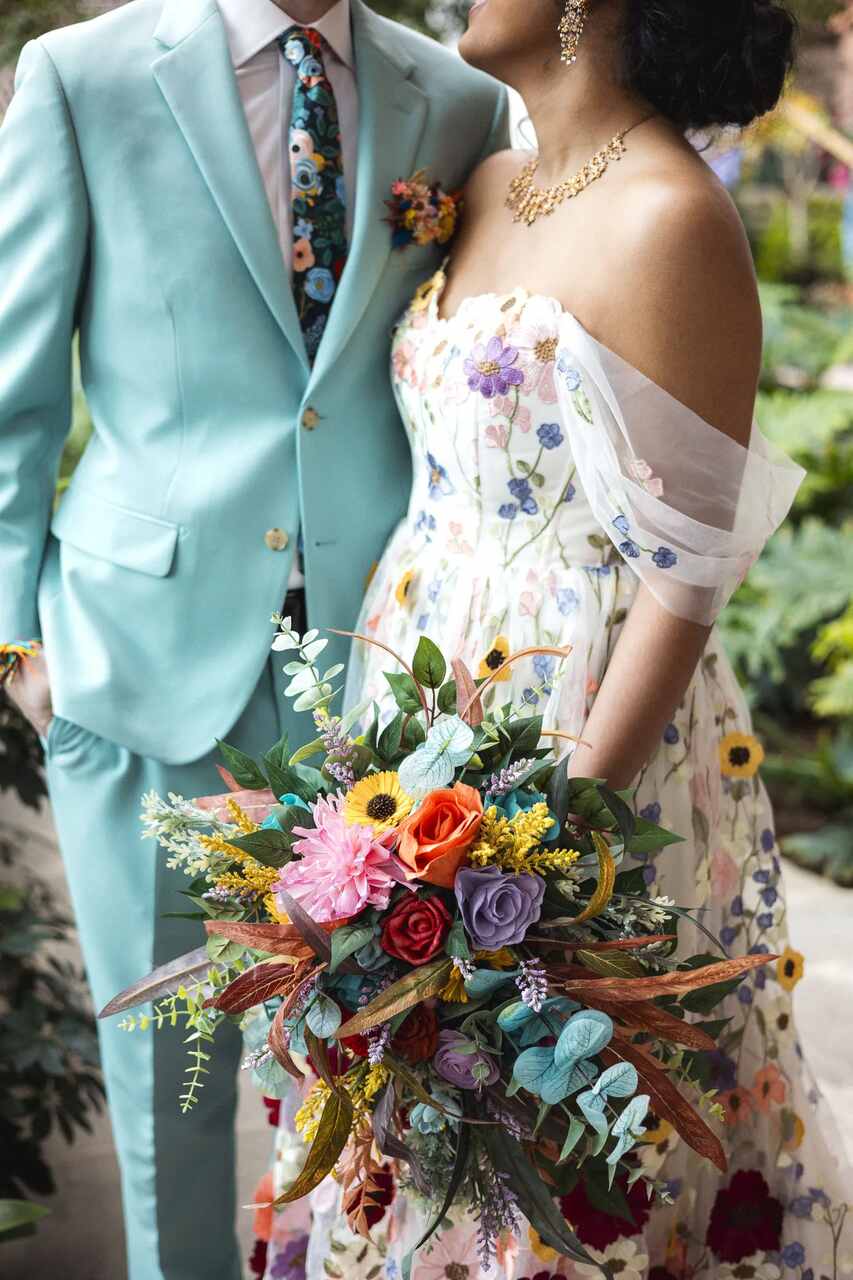 Casal com trajes em tons pastel de casamento; ele com um terno azul-claro e ela com um vestido floral adornado de flores, segurando um buquê colorido.