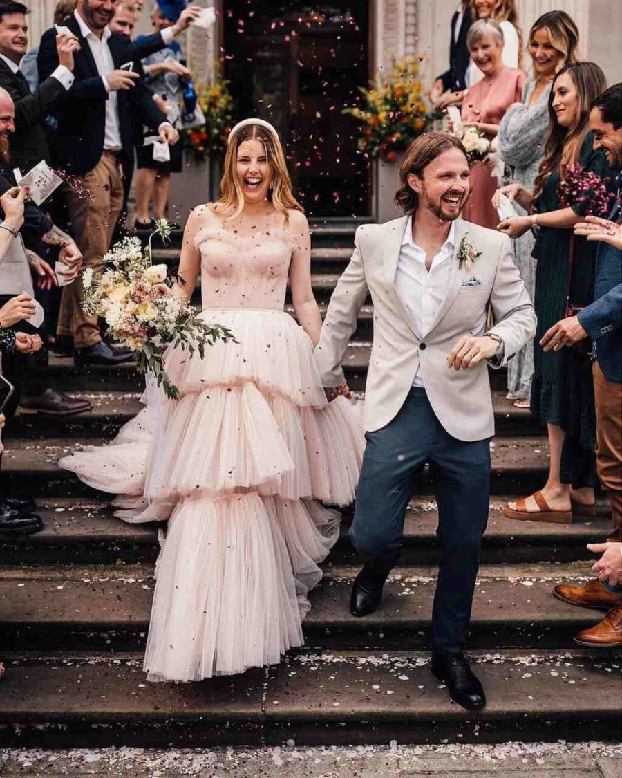 Casal de noivos sorrindo enquanto caminham por uma escadaria, rodeados por convidados jogando pétalas. A noiva usa um vestido volumoso cor de rosa claro com camadas de tule, e o noivo veste um terno claro descontraído.