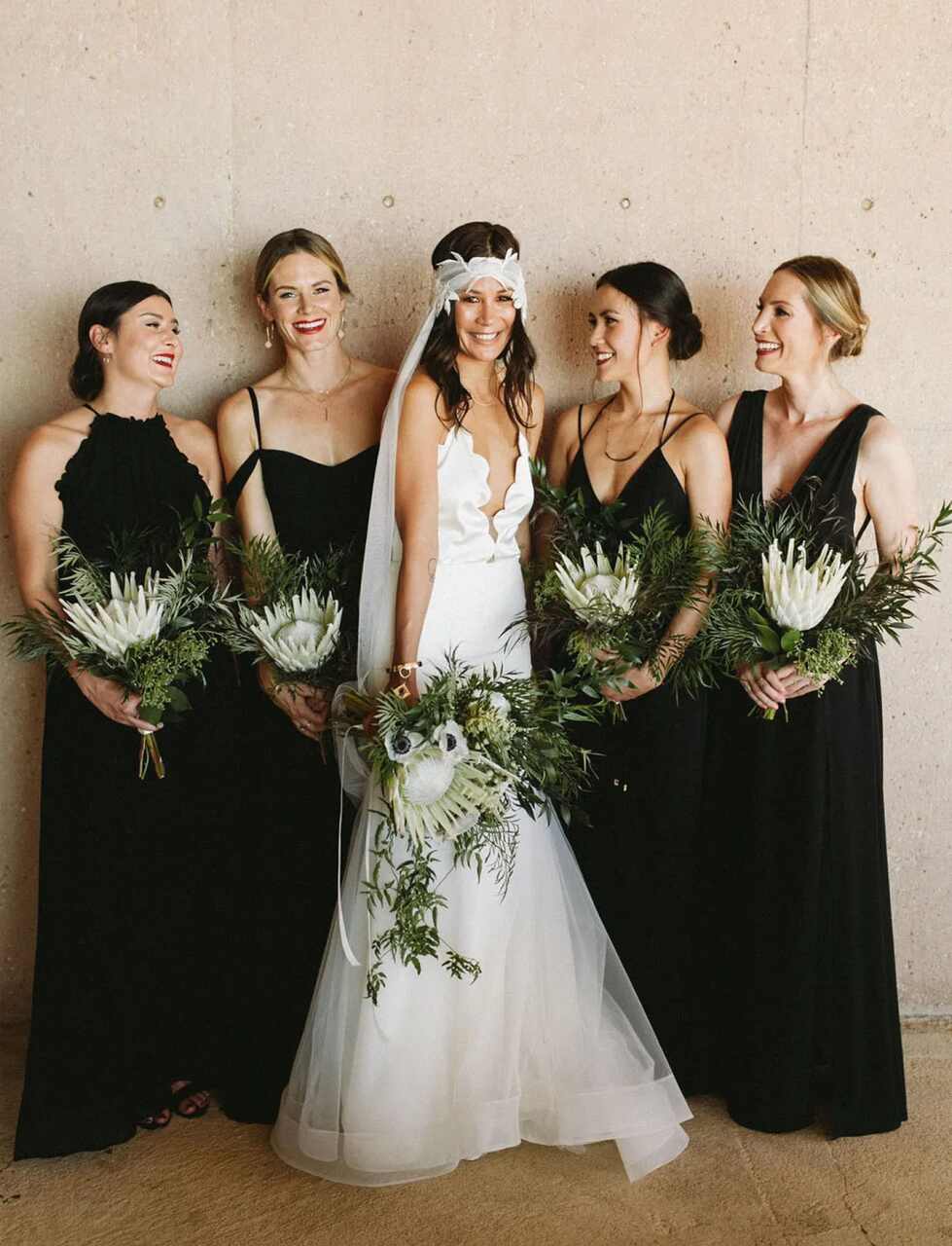 Noiva em vestido branco com detalhes boho, acompanhada por madrinhas de preto segurando buquês de proteas brancas e folhagens exuberantes