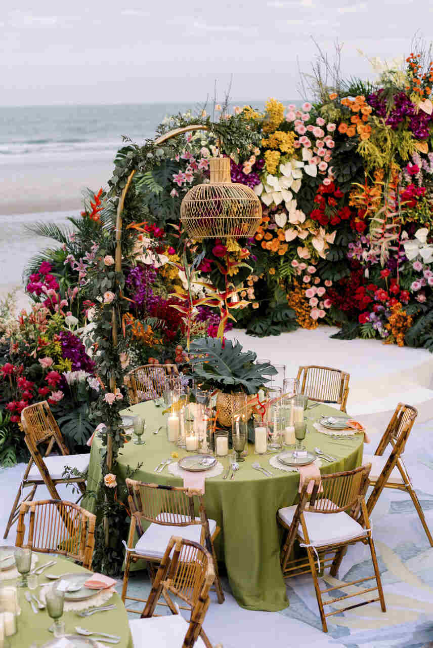 Mesa de recepção em casamento tropical, decorada com toalha verde, flores coloridas, velas e luminária rústica, ao lado de um fundo com vista para o mar