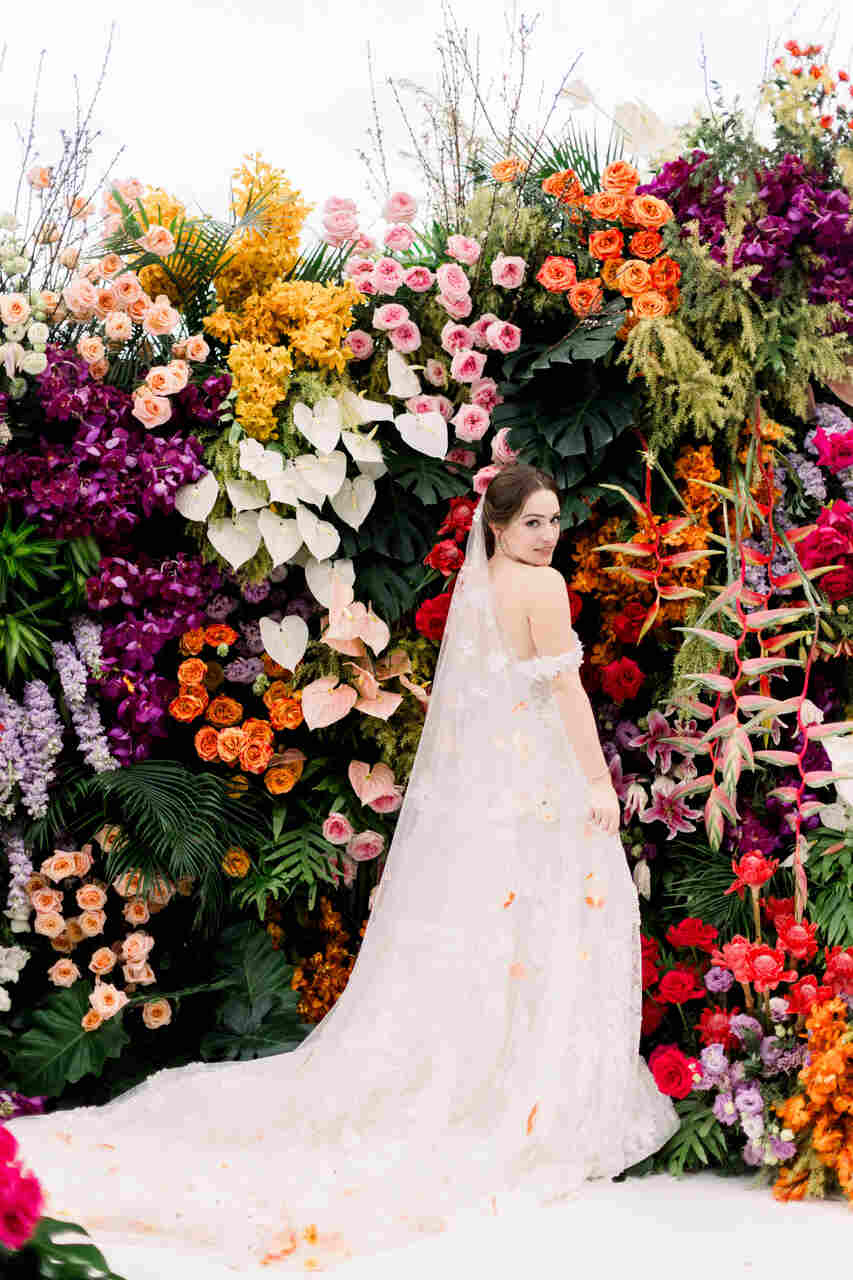 Noiva com vestido branco posando diante de um exuberante arco de flores coloridas.