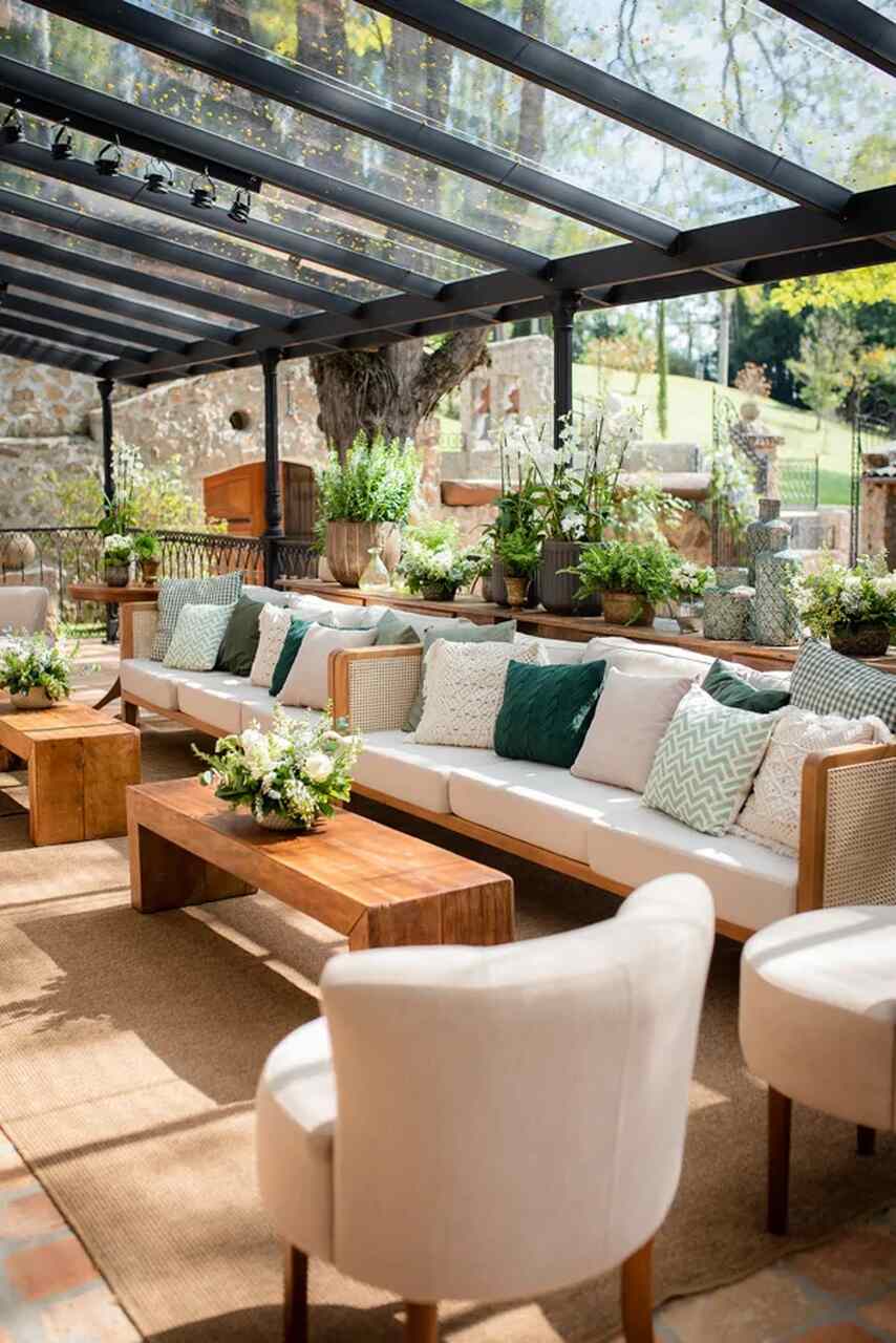 Lounge ao ar livre com sofá branco, almofadas em tons de verde e madeira natural, complementado por uma decoração com muitas plantas.