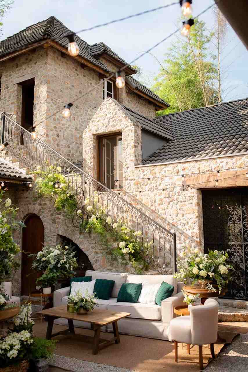 Ambiente rústico e elegante em um casarão de pedra, com sofá branco, detalhes em verde e flores brancas em arranjos