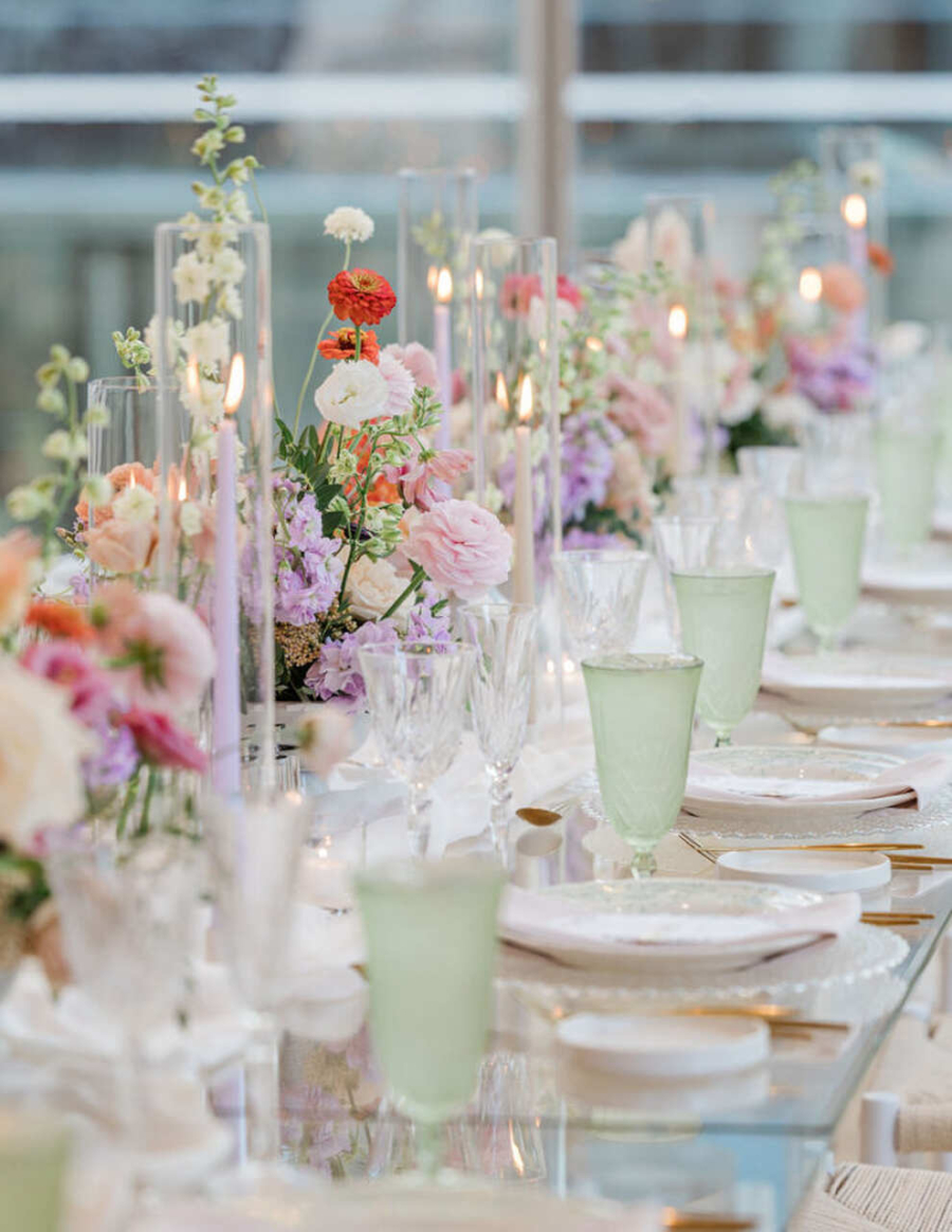 Uma mesa decorada com arranjos florais em tons pastel, velas altas e copos verdes, criando um ambiente iluminado e romântico.