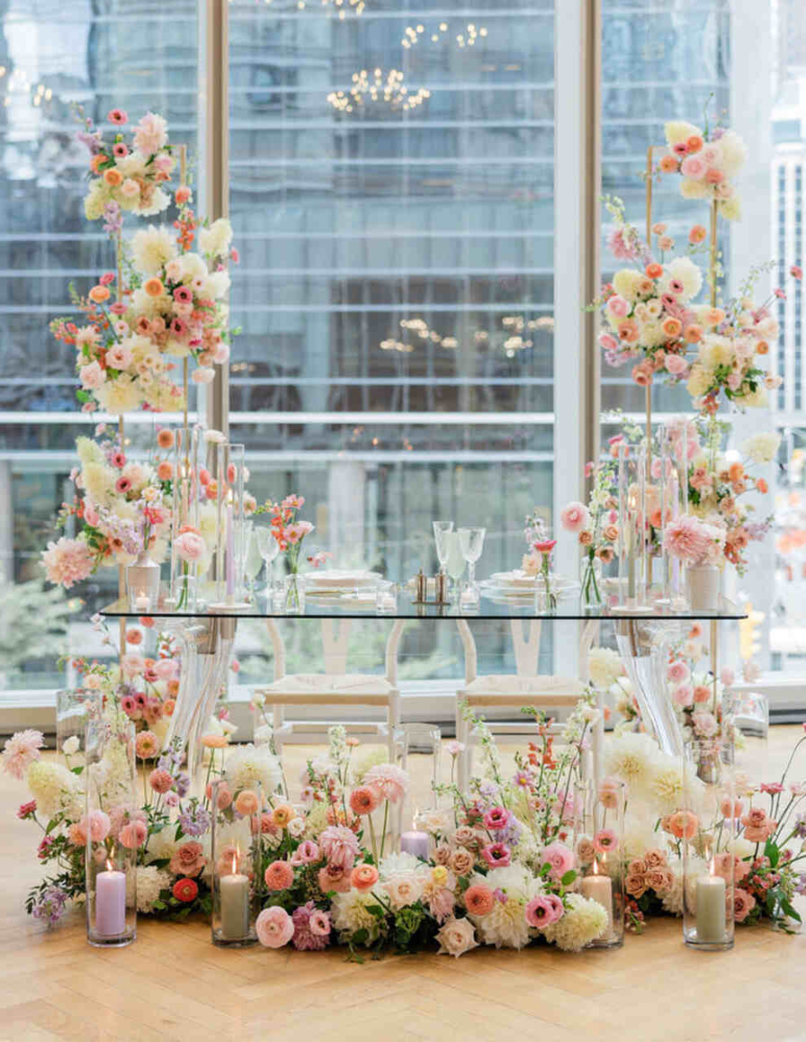 Uma elegante mesa de doces com estrutura de vidro e flores em tons suaves, cercada por velas em um cenário urbano moderno.