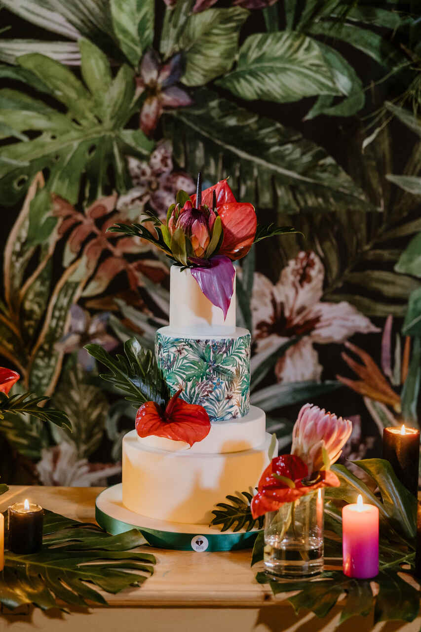 Bolo temático tropical com camadas decoradas por folhagens e flores exóticas, acompanhado por velas coloridas