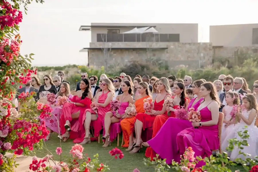 Mulheres sentadas em uma cerimônia ao ar livre, vestindo tons de rosa e laranja vibrantes, com buquês de flores delicados, emolduradas por uma decoração floral exuberante.