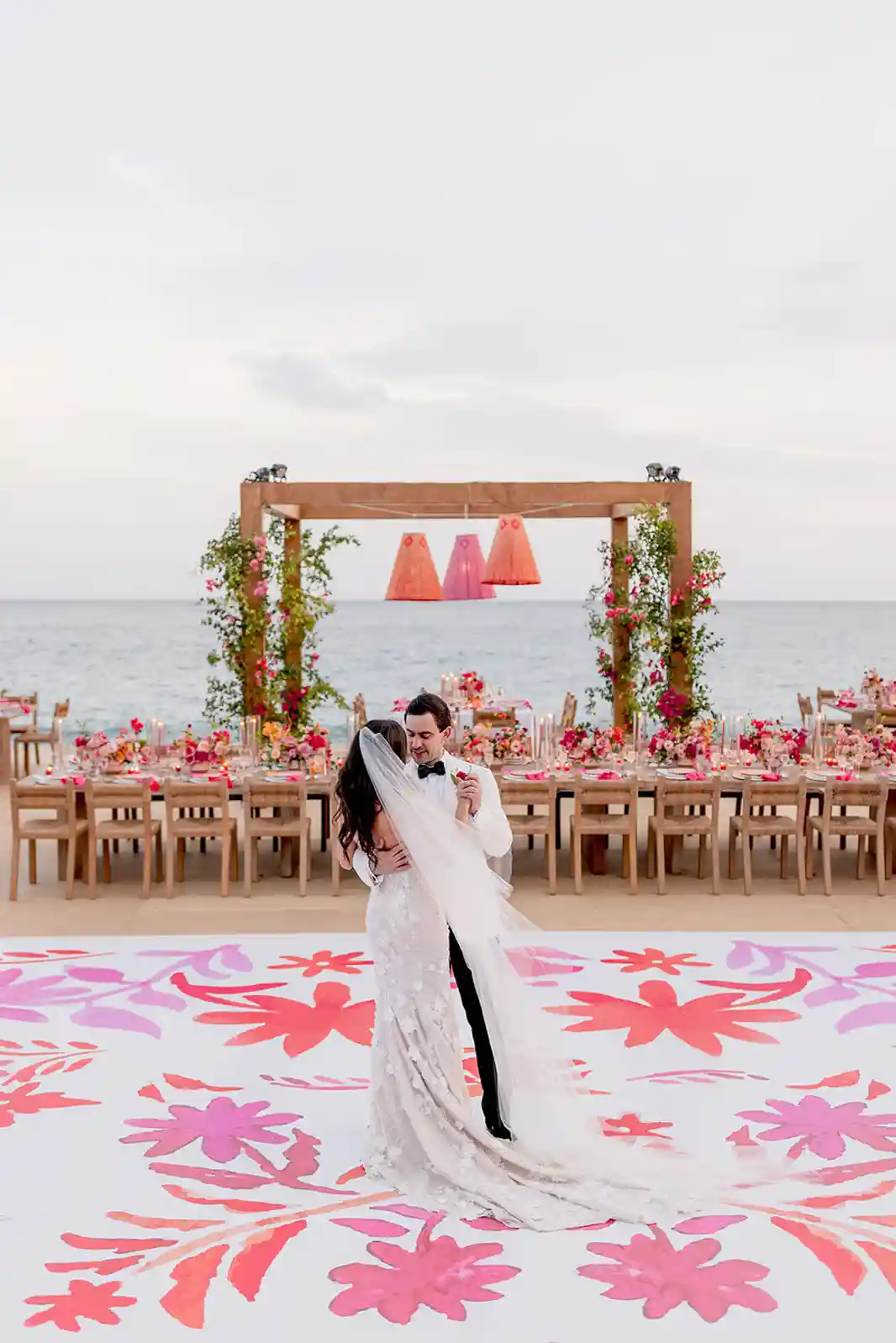 Casal dançando em um casamento tropical ao ar livre, com decoração vibrante em tons de rosa e laranja, flores tropicais, mesas de madeira e vista para o mar.