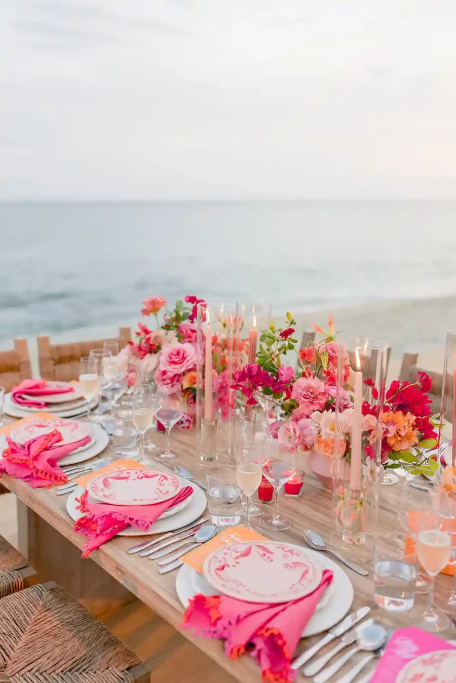 Mesa de jantar à beira-mar decorada com arranjos florais em tons de rosa e velas altas para um jantar elegante ao pôr do sol.