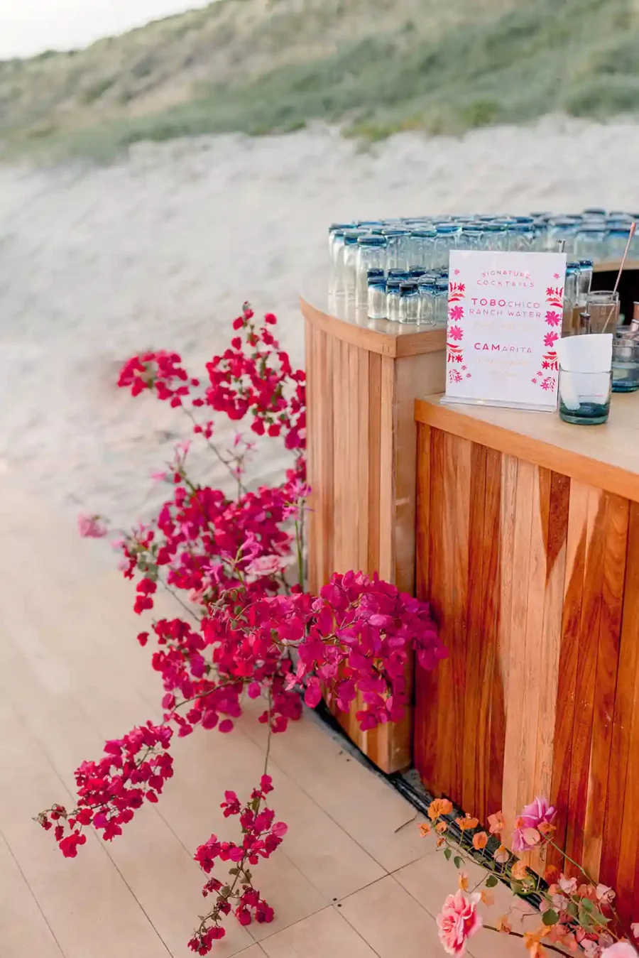 Estação de bebidas com detalhes em madeira, decorada com flores de buganvílias vibrantes, posicionada em uma praia