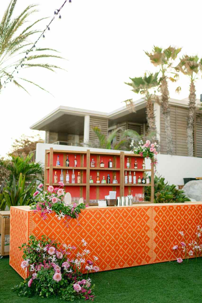 Bar ao ar livre com design laranja vibrante, decorado com flores cor-de-rosa e uma estante de bebidas ao fundo