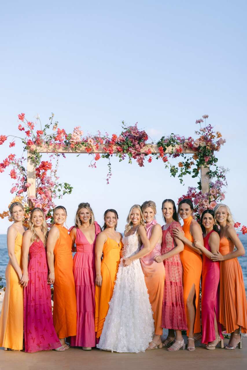 Noiva e madrinhas em vestidos rosa e laranja posando juntas em frente a um arco floral à beira-mar.