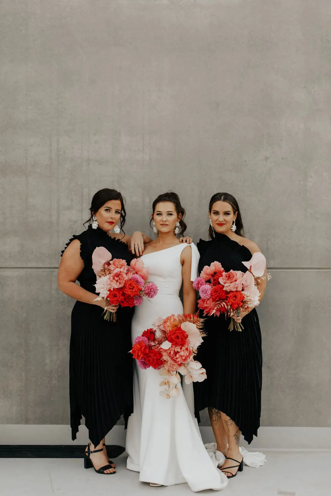 Noiva de branco ao lado de madrinhas em vestidos pretos elegantes, segurando vibrantes buquês de flores em tons de vermelho e rosa.