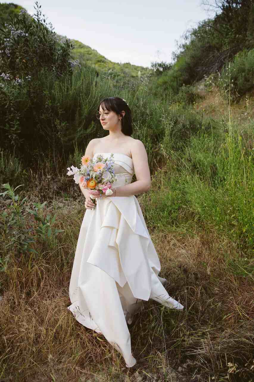 Noiva em um campo sereno, com um vestido de tom claro e design assimétrico, segurando um buquê delicado de flores silvestres.