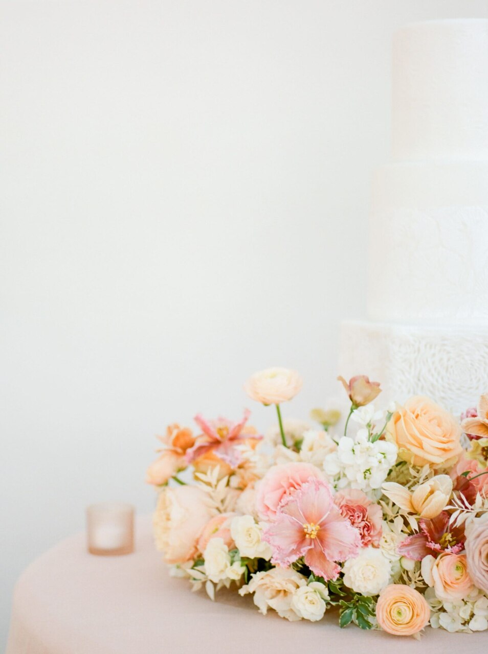 Um bolo branco de casamento, minimalista e elegante, acompanhado por arranjos florais em tons suaves de rosa, pêssego e creme