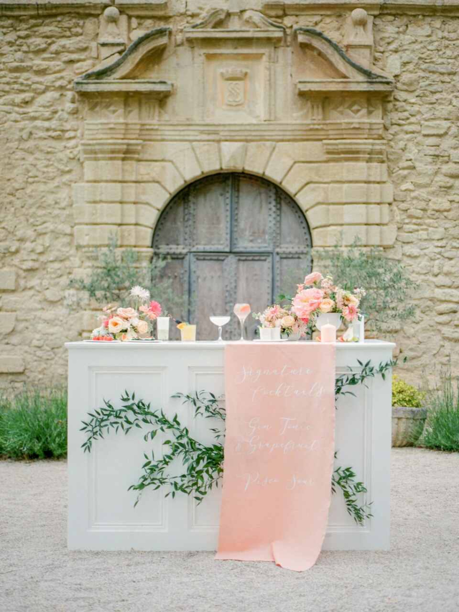 Um balcão branco decorativo posicionado em frente a uma construção de pedra com uma grande porta rústica ao fundo. O balcão está decorado com arranjos florais em tons de rosa, coral e branco, além de folhagens verdes. Uma faixa de tecido rosa pendurada no centro do balcão exibe, em caligrafia delicada, os nomes de coquetéis exclusivos. Ramos de folhas verdes adornam a lateral do balcão, acrescentando um toque natural e elegante à cena. A área ao redor é composta por um chão de pedra clara, complementando o ambiente rústico e sofisticado.