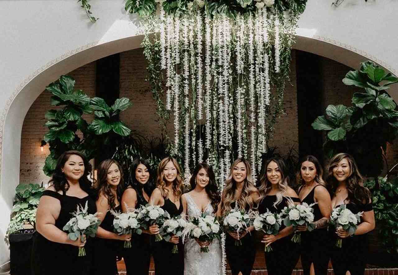 Madrinhas vestidas de preto ao lado da noiva, em um ambiente elegante com decoração de plantas e arranjos pendentes ao fundo.