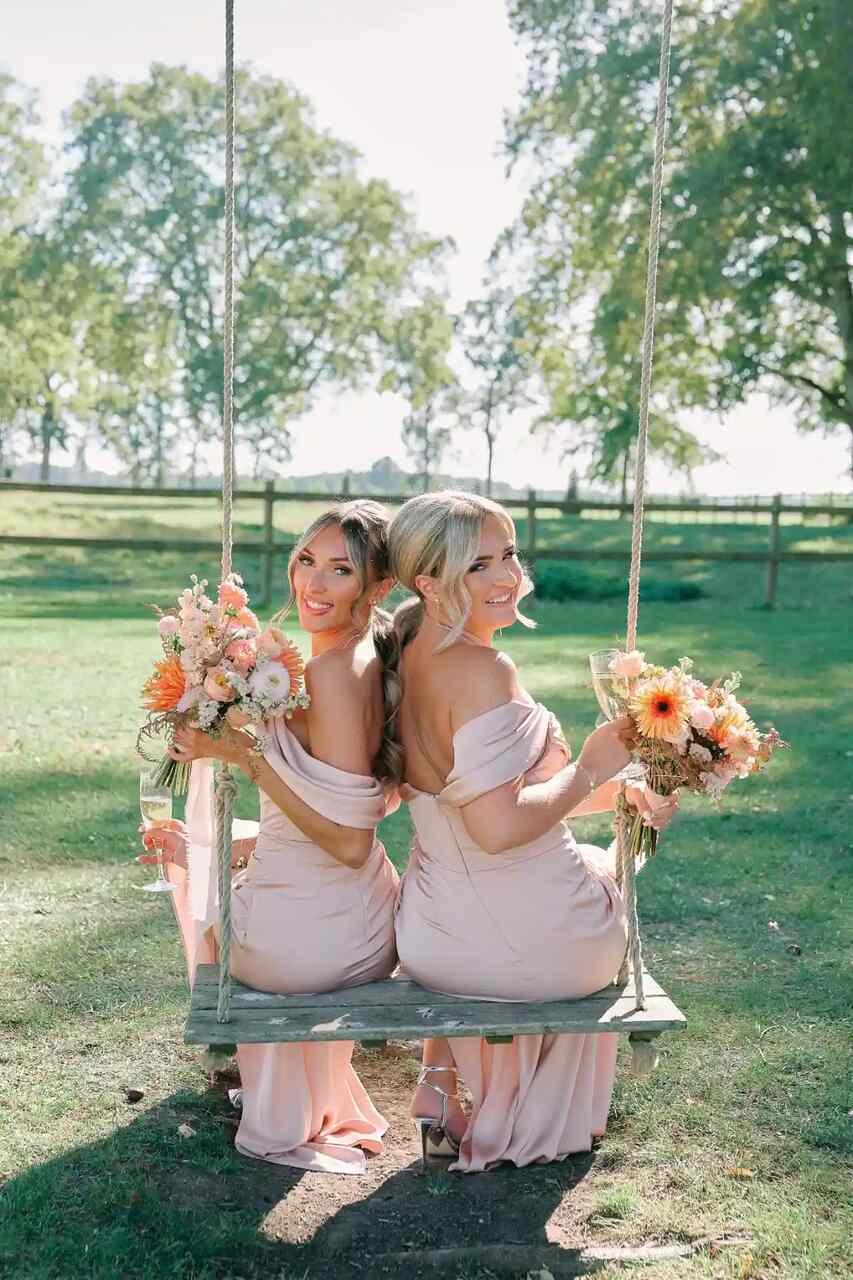 Duas madrinhas vestindo vestidos cor-de-rosa seguram buquês vibrantes enquanto estão sentadas em um balanço ao ar livre.