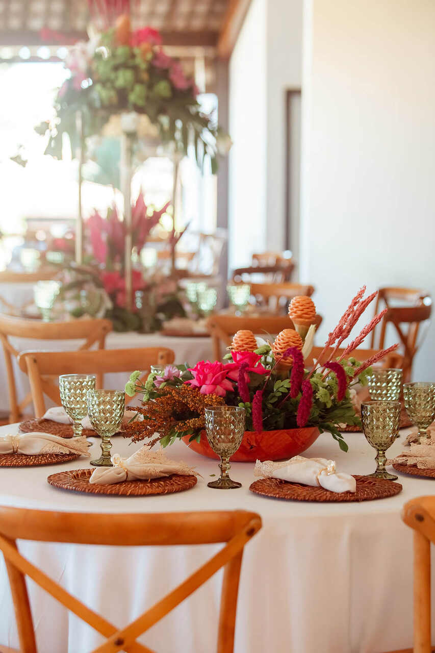 Mesa redonda decorada com arranjo floral tropical e taças verdes, em um salão iluminado com detalhes rústicos