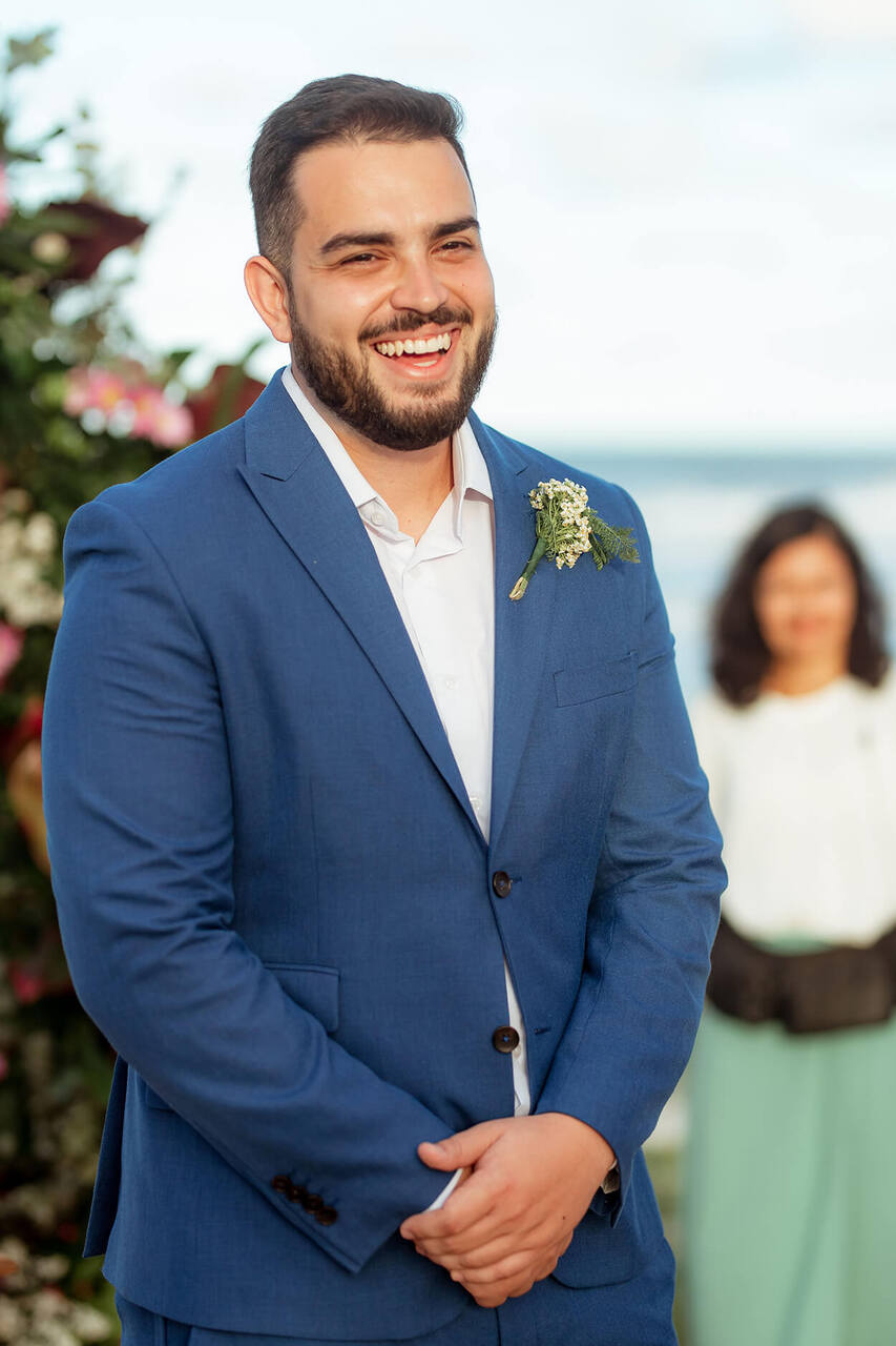 Noivo sorrindo enquanto veste um terno azul e uma lapela floral, em uma cerimônia ao ar livre.