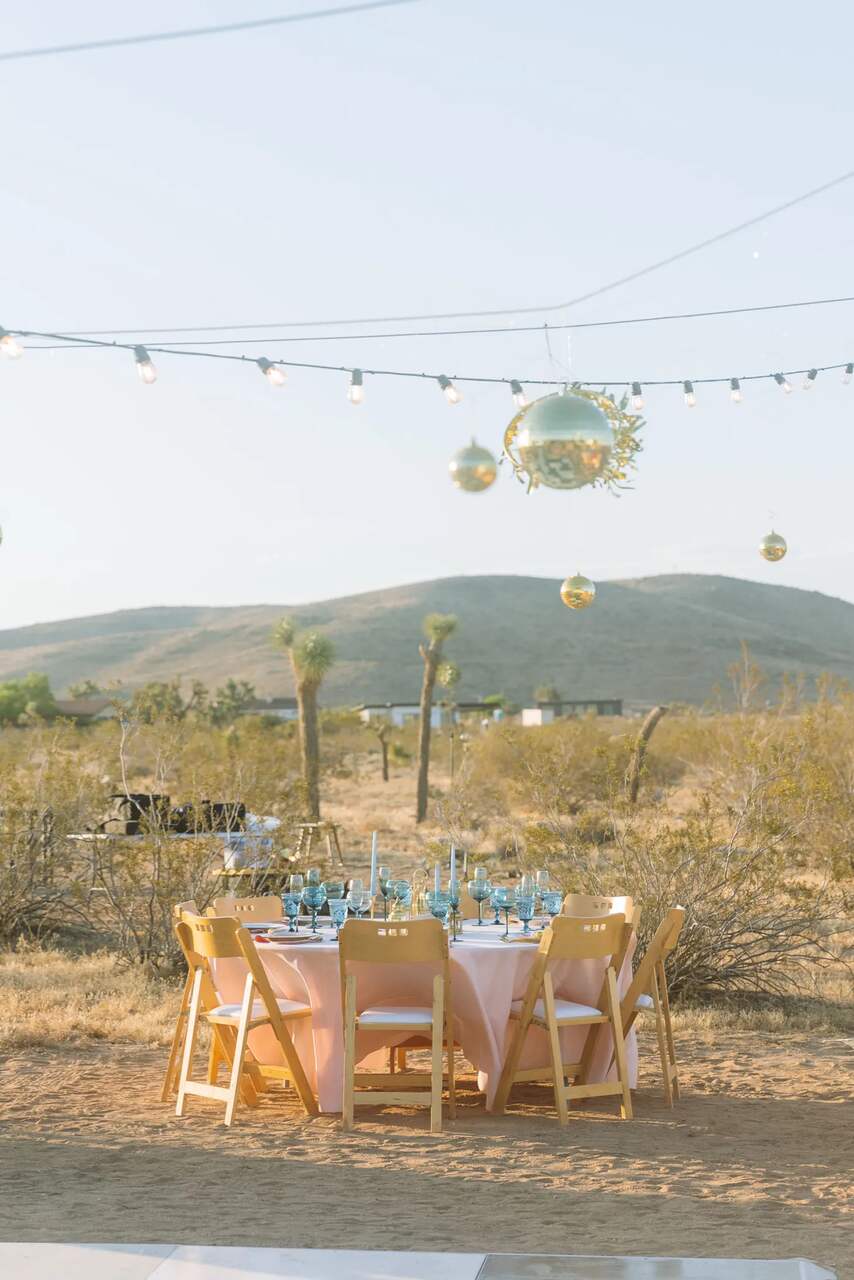 Mesa ao ar livre decorada com louças e copos azuis, cercada por cadeiras de madeira, em um cenário de deserto com luzes e globos dourados suspensos.