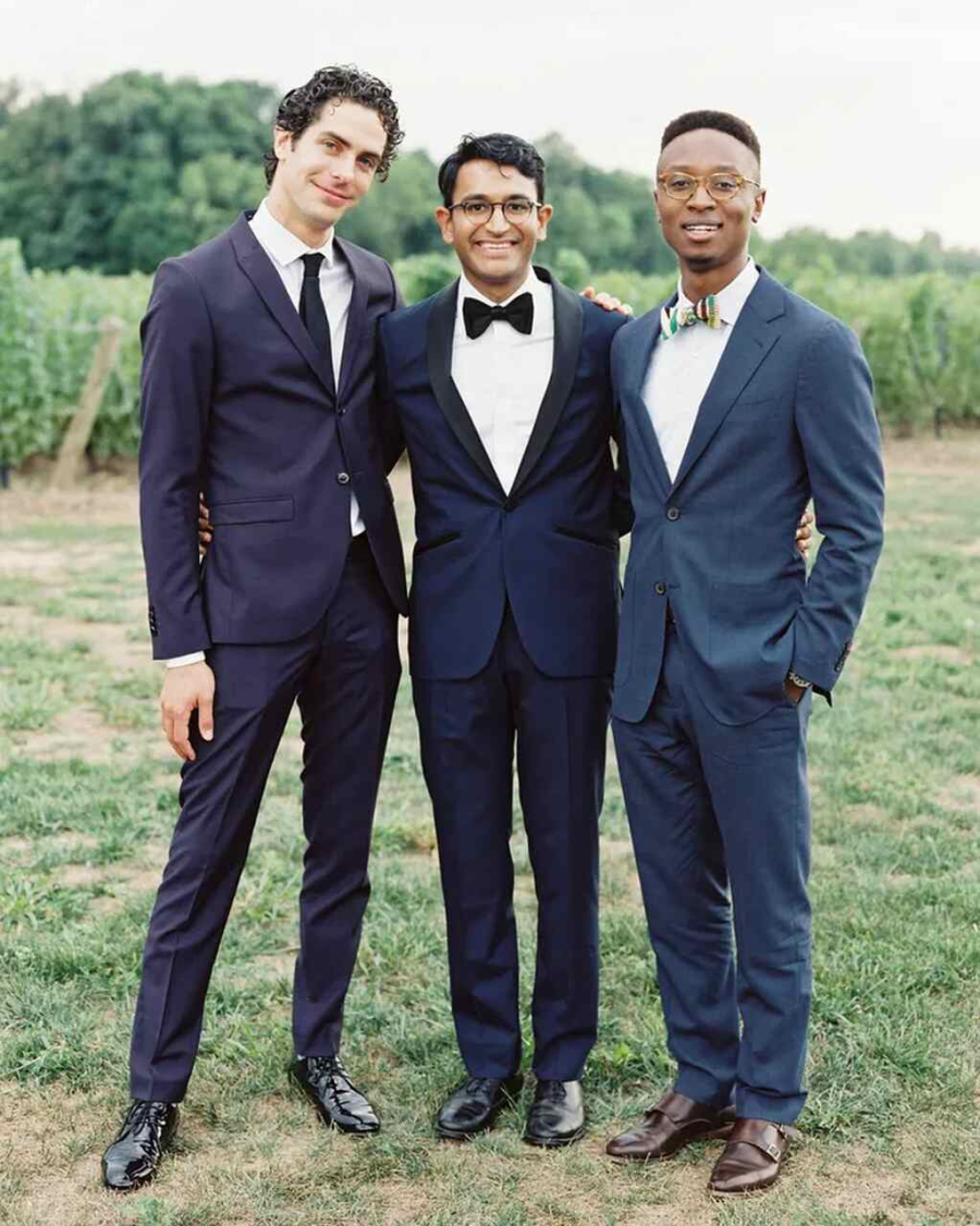 Três amigos posando em um vinhedo, todos de terno; dois com gravatas borboleta e um com gravata clássica.