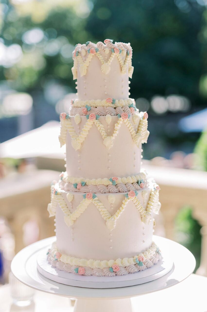 Um bolo de casamento clássico de três andares, decorado com delicadas flores brancas e tons de azul, colocado sobre um pedestal branco em uma mesa decorada com folhagens.