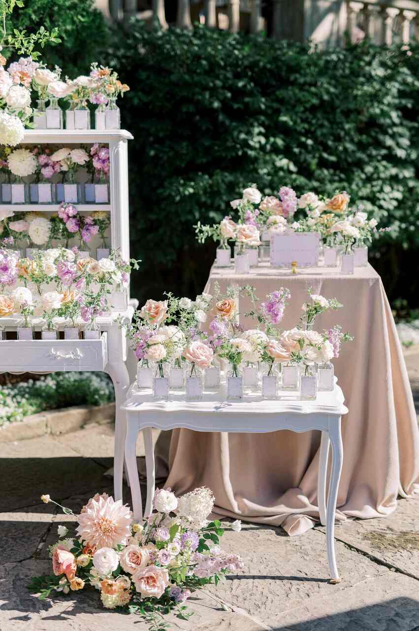 Arranjo delicado de flores em tons pastel dispostos em pequenas garrafas de vidro sobre mesas brancas e forradas, em um jardim iluminado pelo sol.